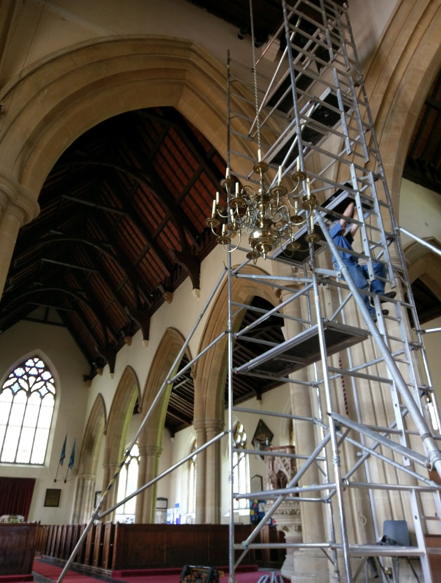An electrician at the top of an access tower, working at ceiling height inside a church beside a brass chandelier