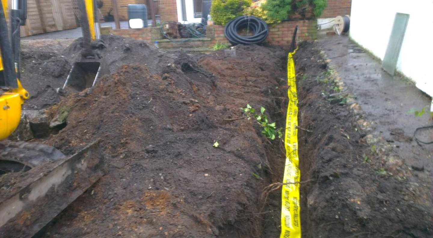 An open trench in a garden with yellow electric cable warning tape laid along it